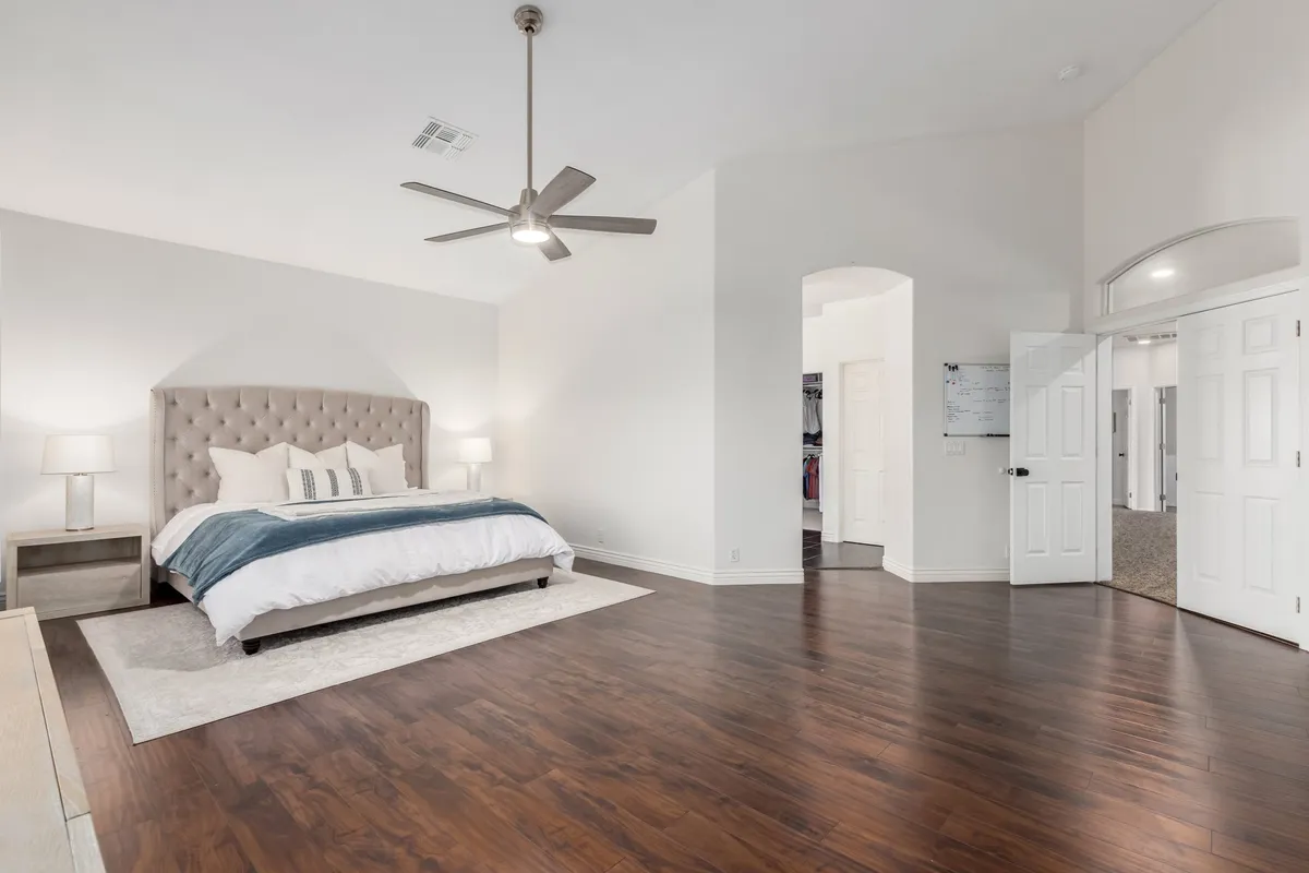 A serene bedroom featuring a king-sized bed with crisp linens, nightstands with matching lamps, a large area rug, and a ceiling fan overhead. The soft lighting and gentle color palette create a peaceful atmosphere, ideal for rest and relaxation.