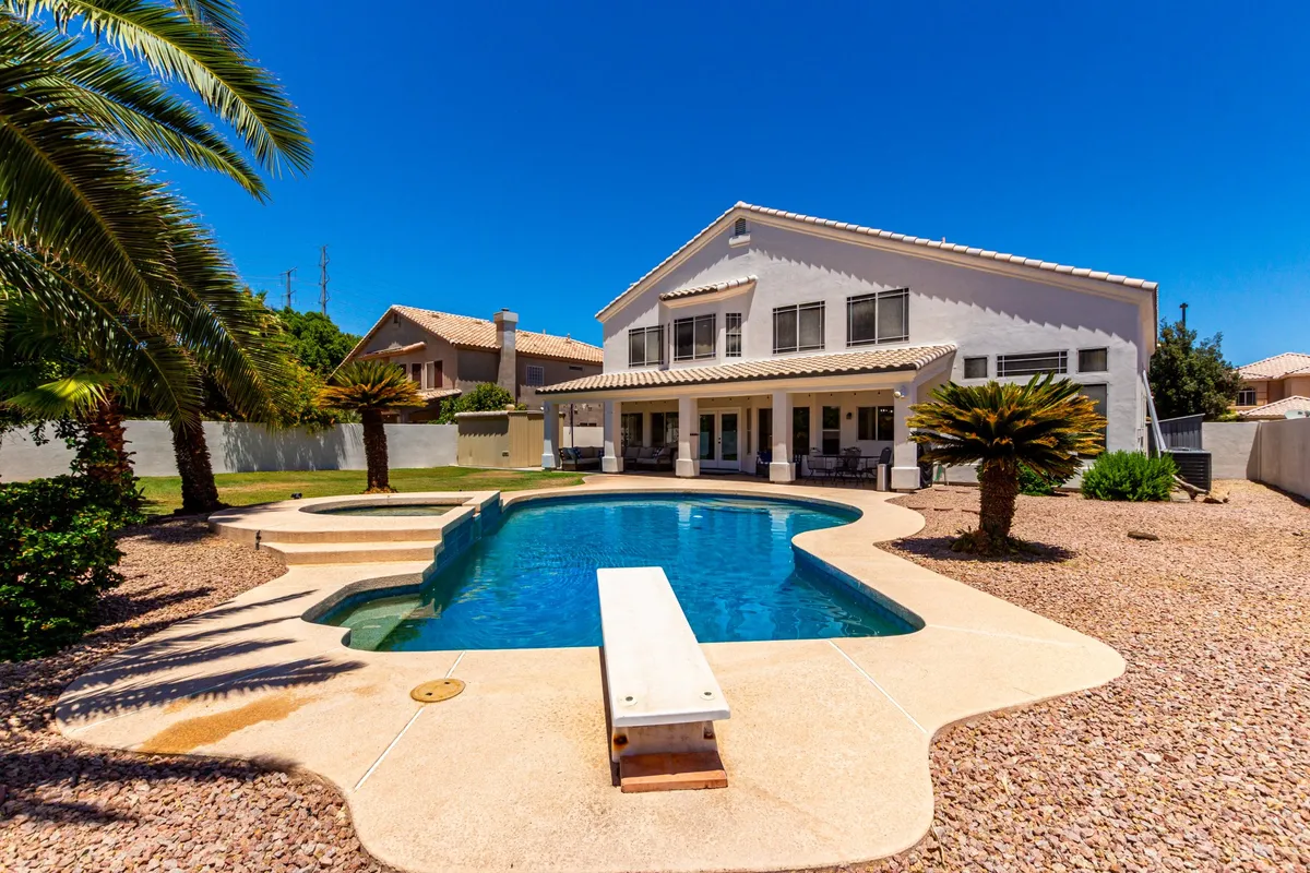 This image showcases a sparkling swimming pool with a dive board, surrounded by palm trees and a view of the house structure. Lounge chairs and shaded areas offer space for relaxation and sunbathing, creating a vibrant, resort-like atmosphere. The scene is lively yet serene, inviting viewers to imagine themselves swimming or enjoying a sunny day by the pool.