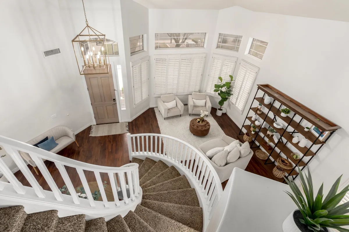A bright, airy space with a spiral staircase, seating area, decorative bookshelf, and abundant natural light from large windows with indoor plants. The atmosphere is inviting and comfortable, ideal for relaxing, socializing, or enjoying a quiet moment after a busy day.
