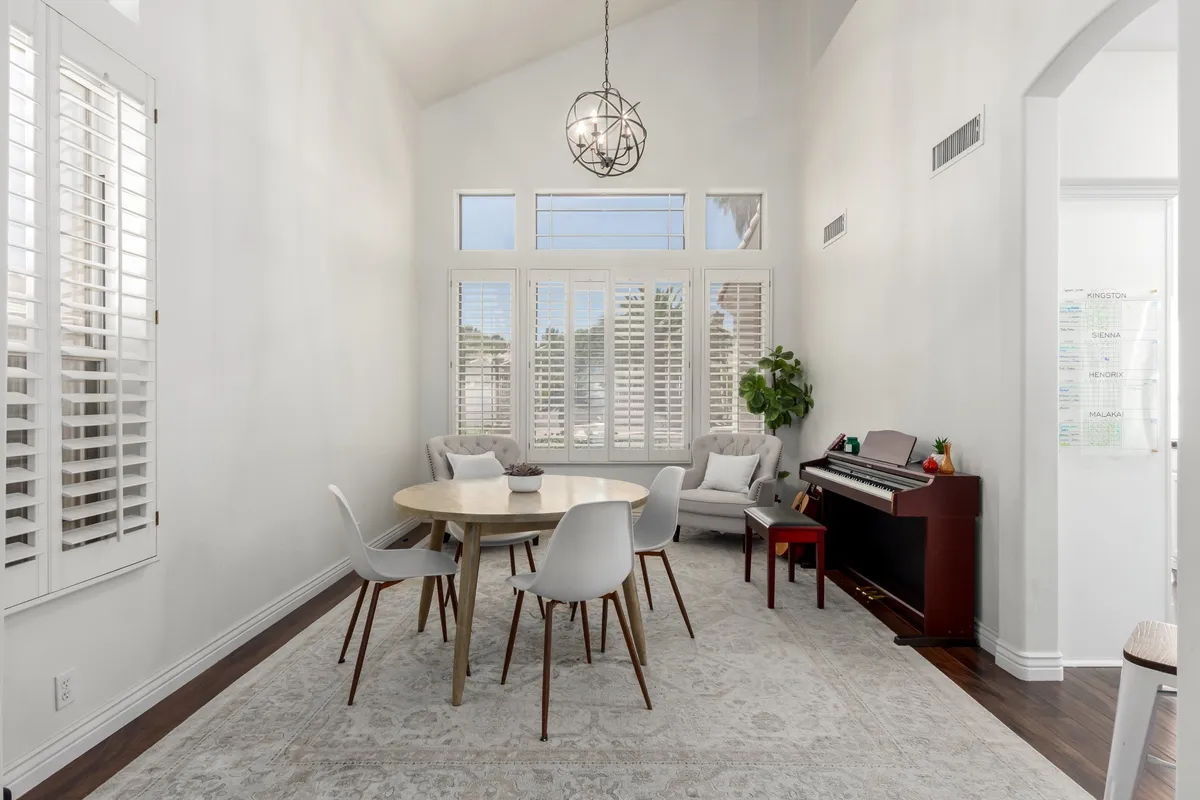 A round wooden dining table surrounded by modern chairs sits near large windows that flood the space with natural light. A piano and lush indoor plant add warmth and a personal touch, creating an inviting setting for meals, music, and casual conversations.