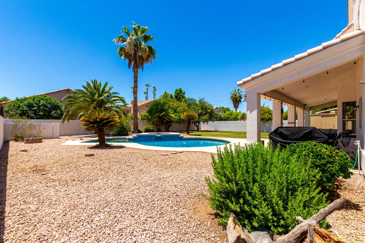 A serene backyard patio area overlooking a sparkling pool, surrounded by palm trees and lush landscaping. The patio space is designed for outdoor dining or lounging, capturing the essence of desert leisure and offering a sense of privacy and calm.