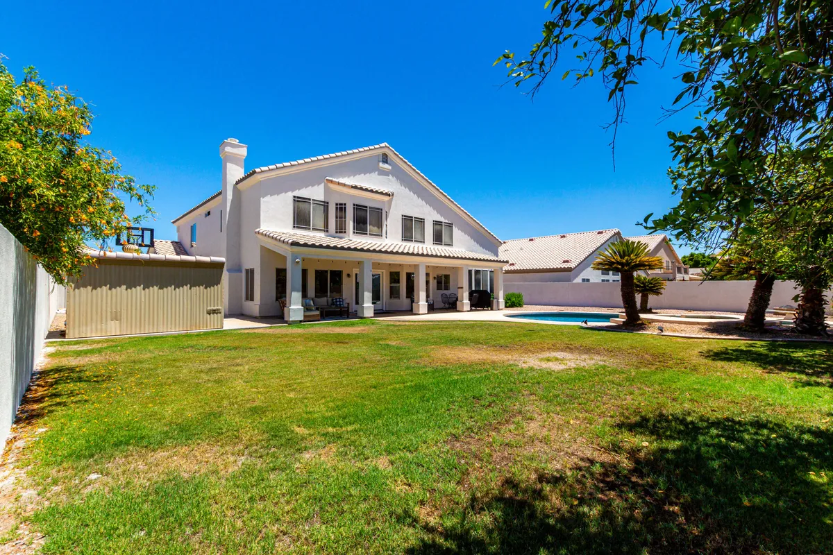 A wide shot of the backyard reveals the swimming pool, patio area, a green lawn, and a basketball hoop. The setup is ideal for both active play and relaxation, with ample space for kids and adults alike. The house facade and clear blue sky frame the fun, energetic vibe of the yard.